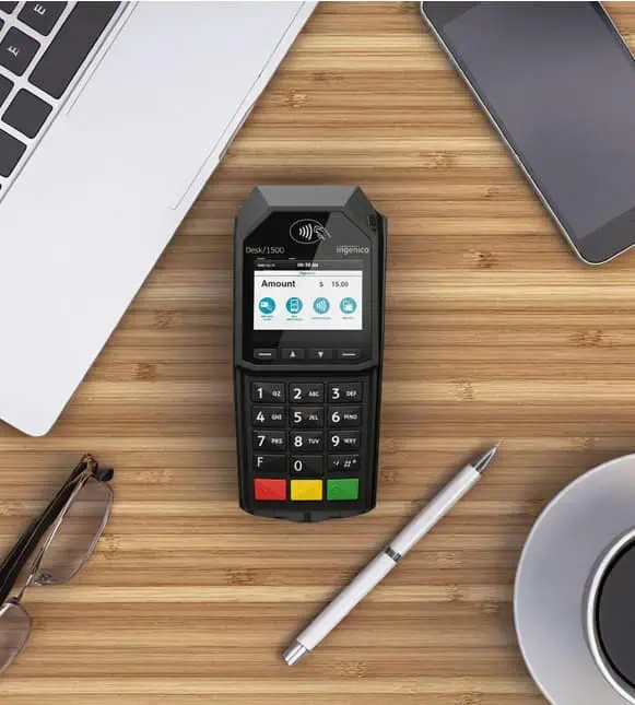 Contactless payment terminal for Host Merchant Services on a wooden desk with laptop and smartphone.